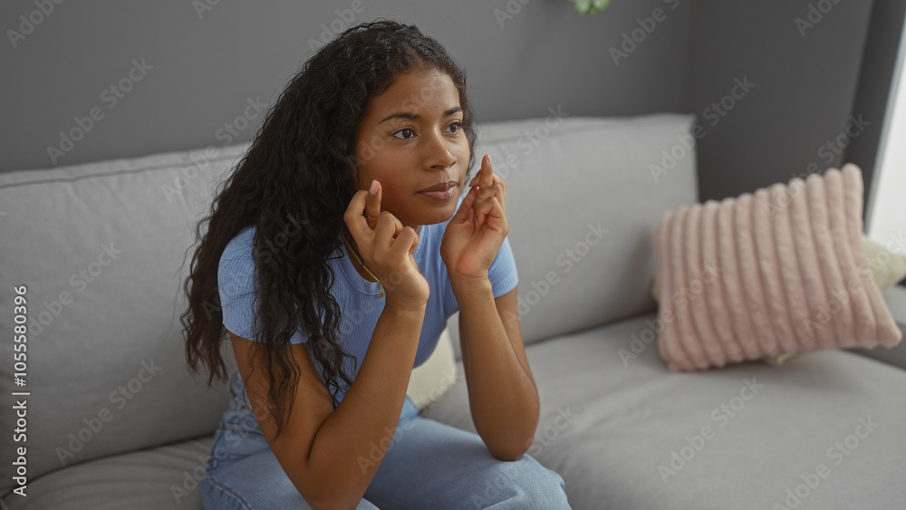 Beautiful young latin woman crosses her fingers while sitting on a couch in her living room, hoping for good luck. Beautiful young latin woman crosses her fingers while sitting on a couch in her living room, hoping for good luck.