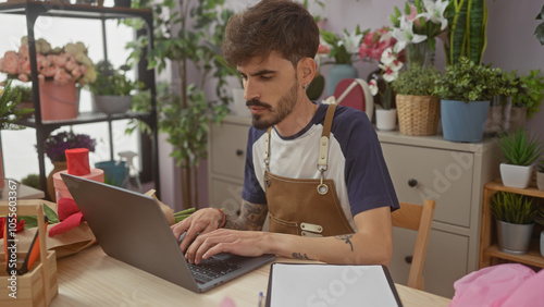 A young hispanic man manages a flower shop, using a laptop surrounded by indoor plants and floral arrangements.