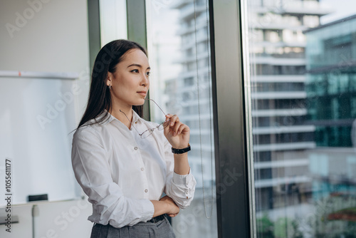 Young businesswoman stands by office window gazing thoughtfully at city skyline