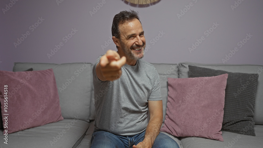 Hispanic man smiling and pointing while sitting on a sofa in a cozy home living room. Hispanic man smiling and pointing while sitting on a sofa in a cozy home living room.