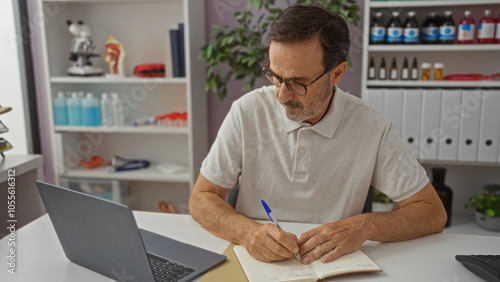 Hispanic man writing at desk in modern pharmacy with laptop and shelves filled with medicinal products, showcasing a mature professional in a clinical store setting.