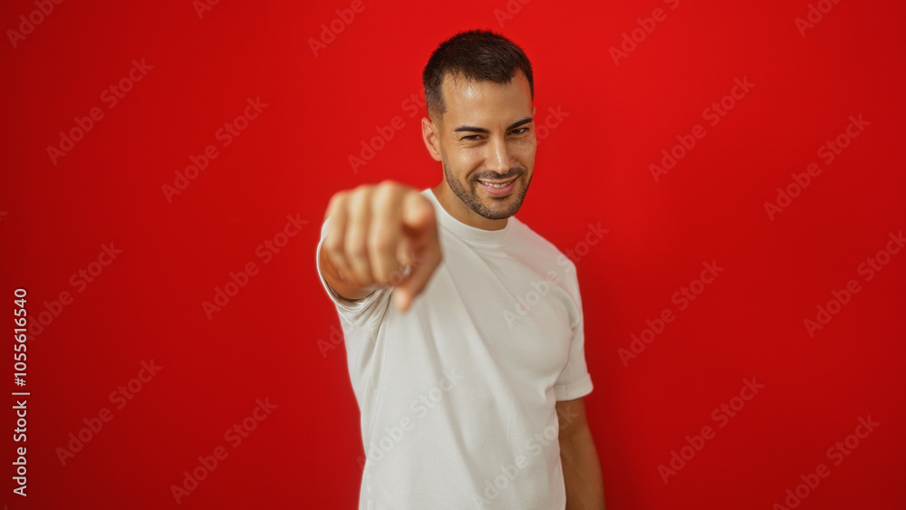 Young hispanic man with a beard in a white shirt pointing at the camera against a vibrant red background Young hispanic man with a beard in a white shirt pointing at the camera against a vibrant red background