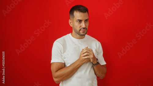 Handsome young hispanic man with a beard standing against a vibrant red background in a white shirt clasping his hands