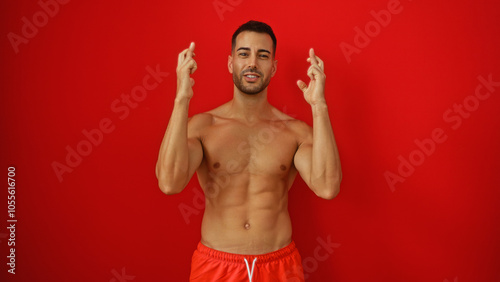 Young hispanic man with a beard standing shirtless against a vibrant red background crossing his fingers