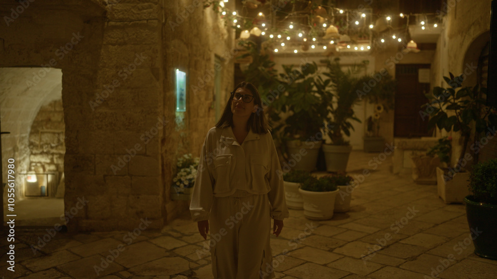Naklejka premium A young hispanic woman walks through the beautifully lit streets of polignano a mare, puglia, italy at night, surrounded by plants and charming ancient stone buildings.