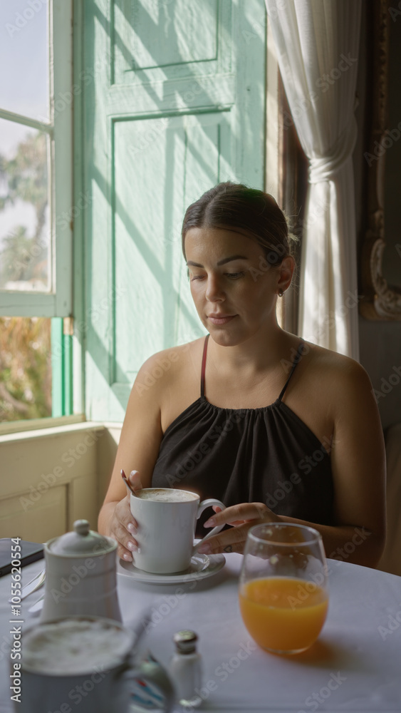 Hispanic woman enjoying coffee indoors at a restaurant table with a window view, morning light highlighting her face and a glass of orange juice on the table. Hispanic woman enjoying coffee indoors at a restaurant table with a window view, morning light highlighting her face and a glass of orange juice on the table.