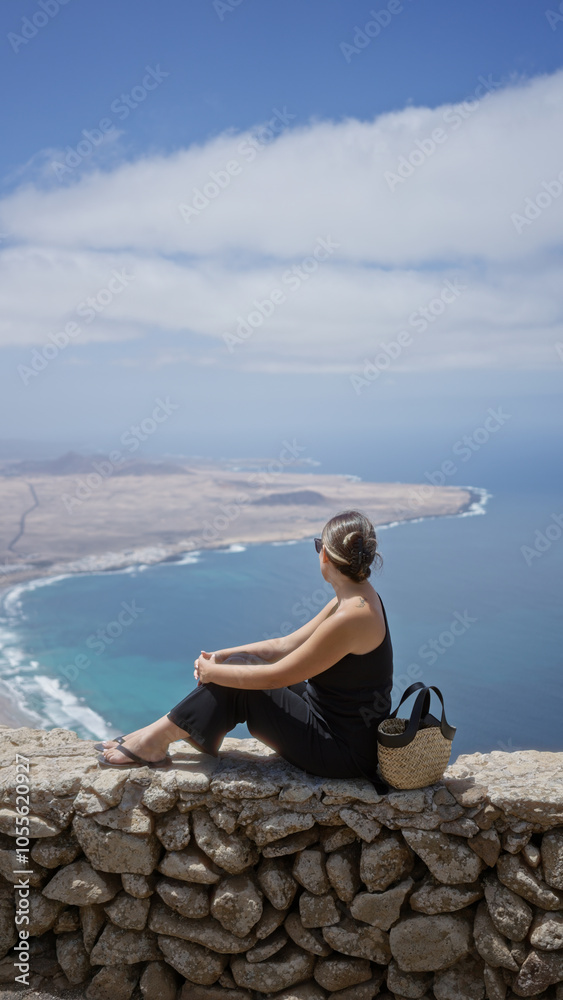 Young woman sightseeing in lanzarote, canary islands, sitting on a stone wall with a view of the coastline and ocean, enjoying a sunny outdoor day. Young woman sightseeing in lanzarote, canary islands, sitting on a stone wall with a view of the coastline and ocean, enjoying a sunny outdoor day.