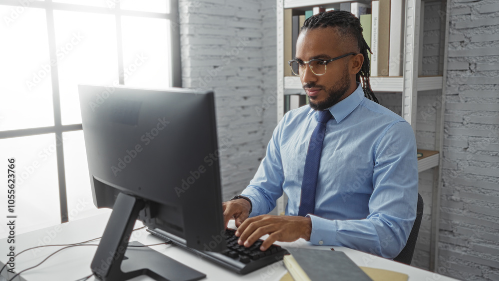Obraz premium Young man working on computer in modern office wearing blue shirt and glasses focused on screen daytime setting with shelves in background suggesting professional environment