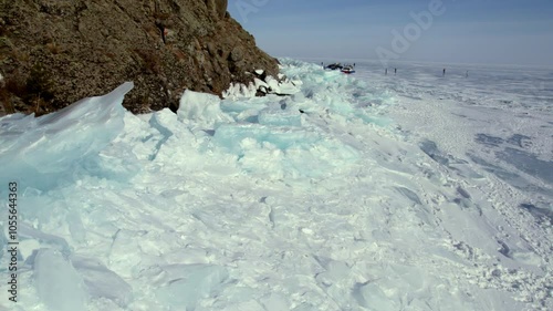 Huge blocks of ice near the rocky shore of the frozen Lake Baikal. Tourists walk on the ice. Hovercraft are waiting for passengers.