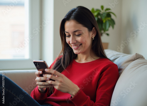 Obraz na płótnie A young woman with long black hair is sitting on a couch and smiling while looking at her phone. She is wearing a red sweater. w salonie