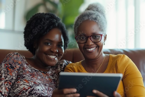 Two women are sitting on a couch and smiling while looking at a tablet. The tablet is being held by one of the women. Concept of warmth and happiness, as the women are enjoying each other's company