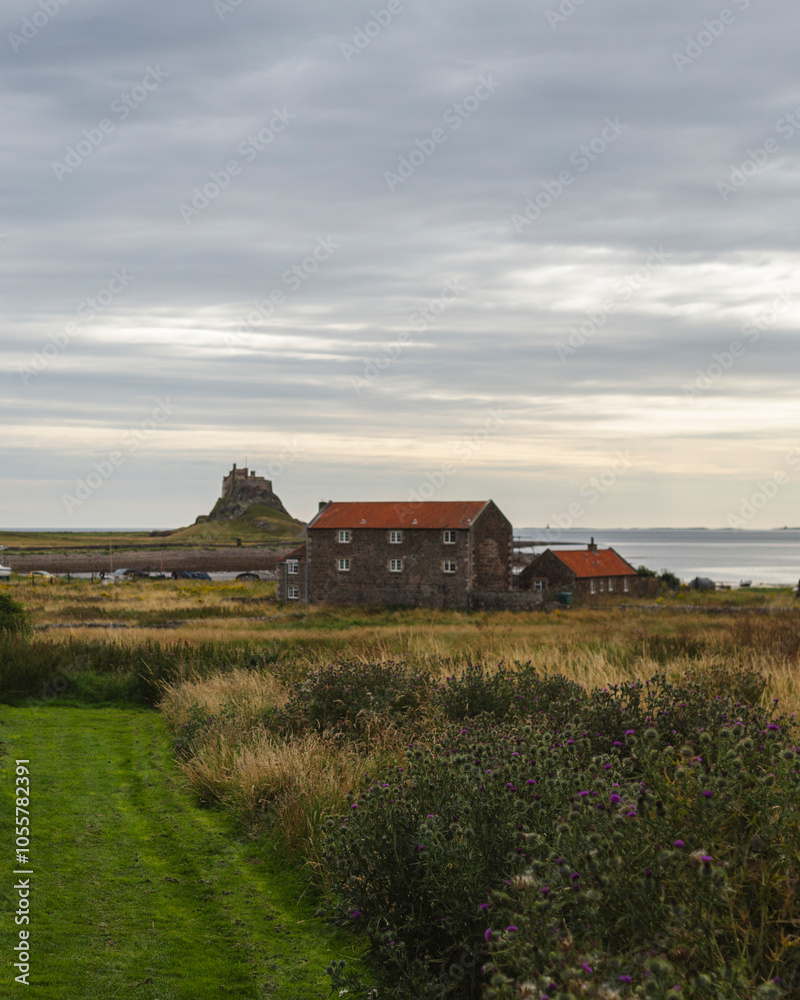 View of historic castle and stone buildings amidst tranquil fields and coastline under a cloudy sky, Holy Island, England. View of historic castle and stone buildings amidst tranquil fields and coastline under a cloudy sky, Holy Island, England.