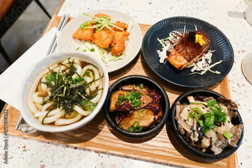 close up of soup udon with seaweed, mushroom appetiser, grilled salmon fish, eggplants and fried chicken in small plates bowls in a wooden tray