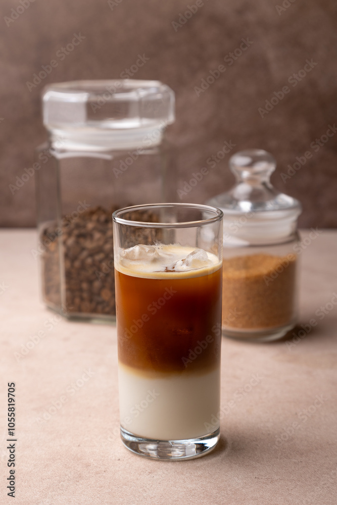 Coffee with coconut milk and ice cubes in a tall glass. Traditional non-alcoholic cold drink. In the background, a jar of coffee and a jar of sugar. Selective focus, close-up. Coffee with coconut milk and ice cubes in a tall glass. Traditional non-alcoholic cold drink. In the background, a jar of coffee and a jar of sugar. Selective focus, close-up.