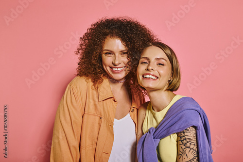 A joyful lesbian couple with natural makeup radiates love and diversity against a pink backdrop.