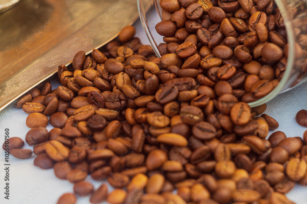 Fototapeta premium A pile of red roasted coffee beans spilled out of an overturned transparent glass jar onto the white surface of the tablecloth on table. One of the most important export products in some countries