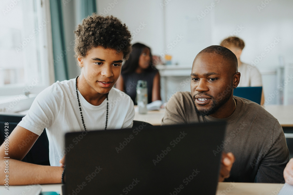 Male student and teacher using laptop at desk in classroom Male student and teacher using laptop at desk in classroom