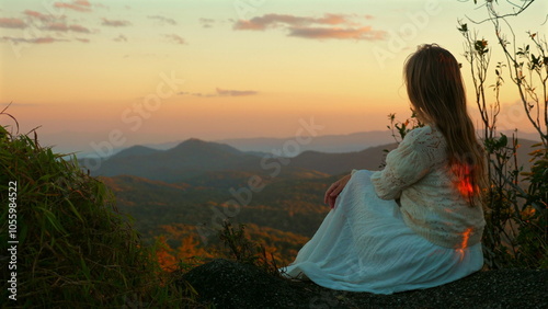 Woman sitting on rock and enjoying serene mountain sunset view, wearing white dress, representing peace and nature appreciation.