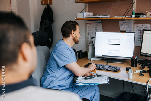 Male doctor checking medical records of patient on computer while sitting near table at medical examination room photo