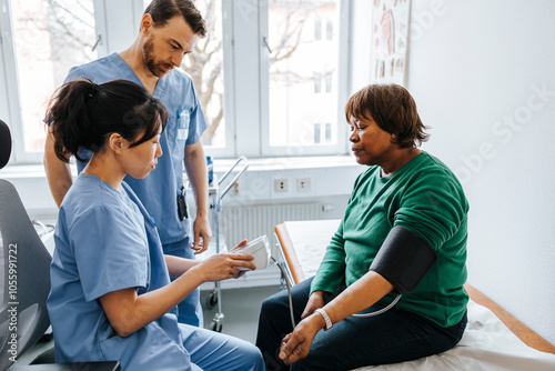 Female doctor checking blood pressure temperature of senior patient on gauge with coworker standing in examination room photo