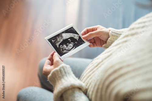 Fotografie Close-up - Hand of happy pregnant woman watching her ultrasound report and touching her abdomen while sitting on bed at home, Maternity love