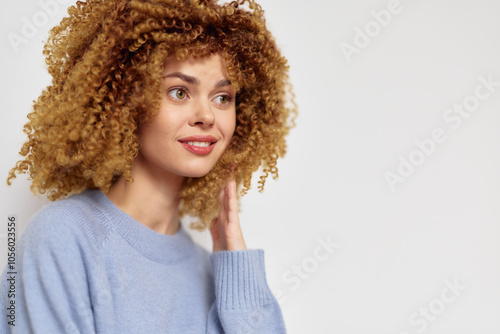 Smiling young woman with curly hair in a cozy blue sweater against a light neutral background, showcasing a warm and inviting expression, perfect for lifestyle concepts