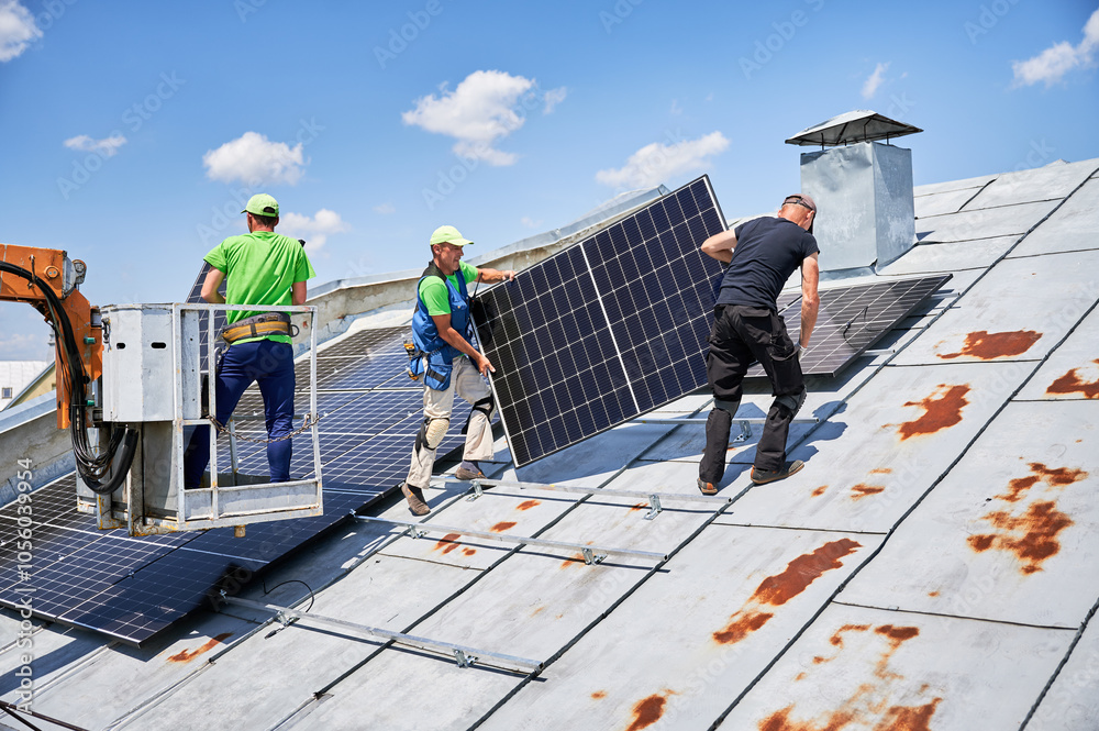 Workers building solar panel system on metal rooftop of house with assistance of crane lift. Two men installers carrying photovoltaic solar module outdoors. Renewable energy generation concept. Workers building solar panel system on metal rooftop of house with assistance of crane lift. Two men installers carrying photovoltaic solar module outdoors. Renewable energy generation concept.