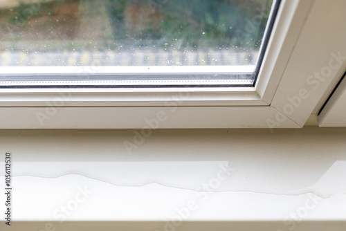 Top view of a white window frame with a window sill, which spreads a puddle. There is a gap in the double-glazed window, the window frame is leaking