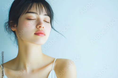 Portrait of a serene woman with closed eyes, soft makeup, and relaxed expression, illuminated by gentle, natural light against a pale background. Copy space on right.