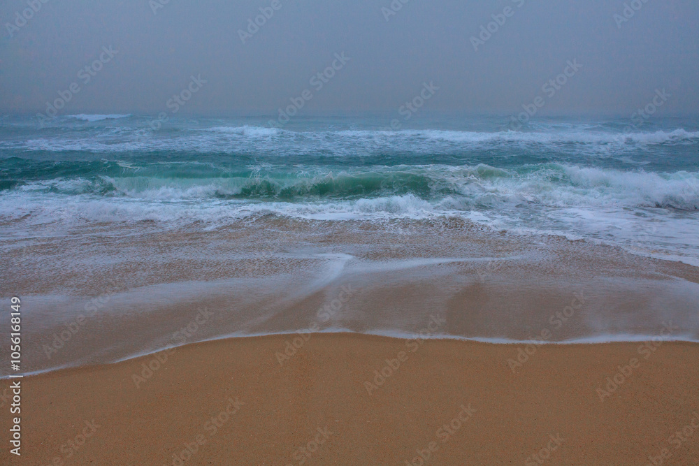 Gentle waves roll towards the sandy shore under a misty, overcas Gentle waves roll towards the sandy shore under a misty, overcas