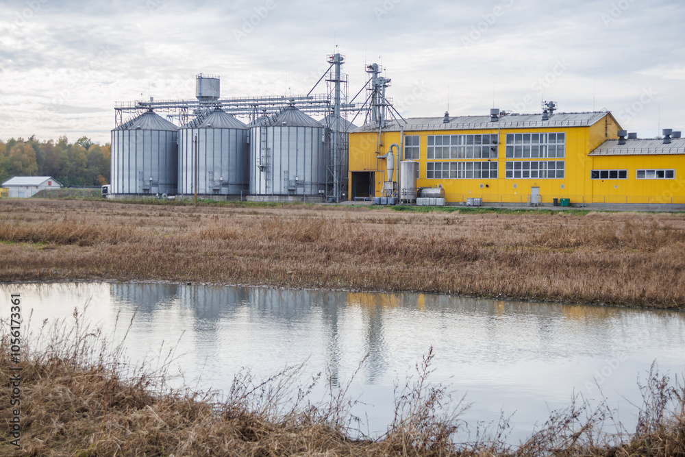 large grain elevator with multiple cylindrical metal silos, structural elements, and a complex system of pipes. It stands against a blue sky, with a field in the foreground after harvest large grain elevator with multiple cylindrical metal silos, structural elements, and a complex system of pipes. It stands against a blue sky, with a field in the foreground after harvest
