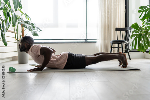 Young African-American man doing yoga on a mat in a bright home setting, practicing the cobra pose with focused posture. Surrounded by plants, promoting wellness and relaxation.