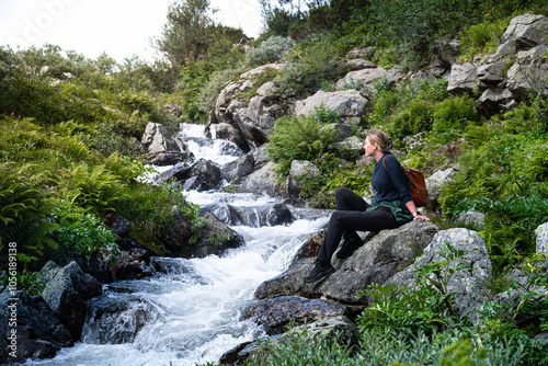 Woman sitting peaceful among rocks, gazing at the cascading Silverfallet waterfall as bright sunlight shining on her face in Dalarna Sweden, surrounded by lush green and the soothing sound of water