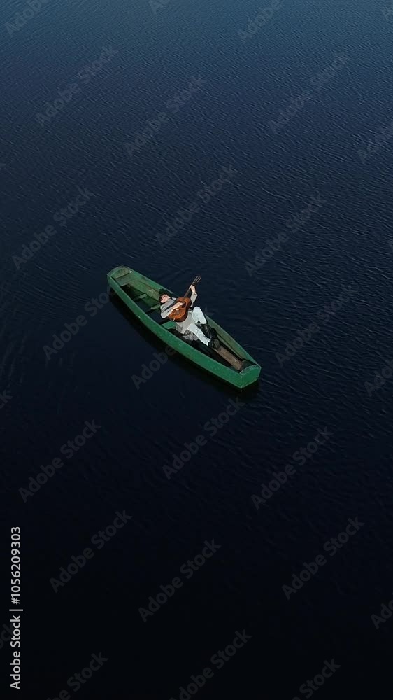 Vertical video. A man plays the guitar lying in a wooden boat, in the middle of a lake on the water in the middle of nature. Aerial view. Vertical video. A man plays the guitar lying in a wooden boat, in the middle of a lake on the water in the middle of nature. Aerial view.