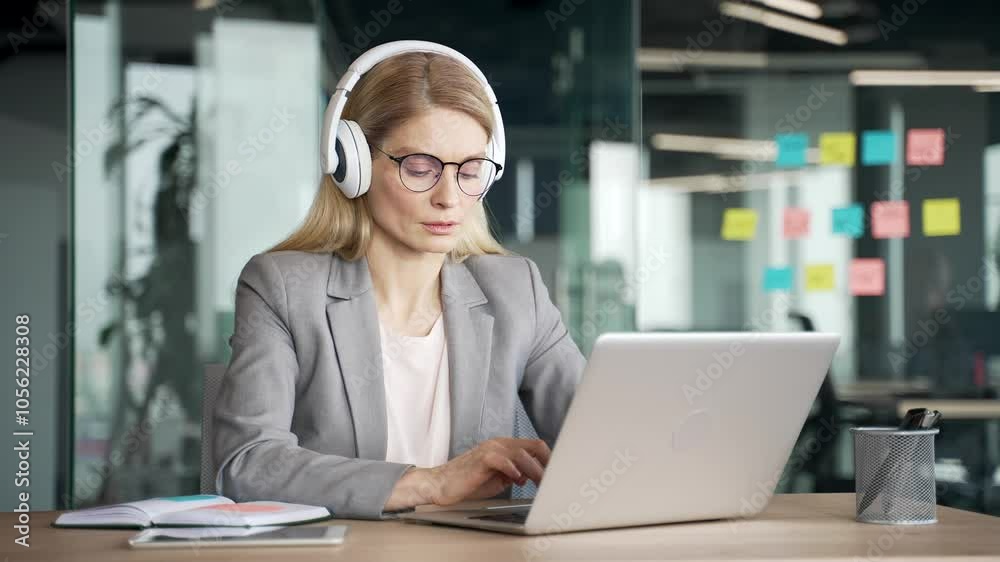 Confident businesswoman in headphones typing on laptop sitting at workplace in business office. Serious female IT specialist in a suit wearing glasses works on computer while listening to podcast