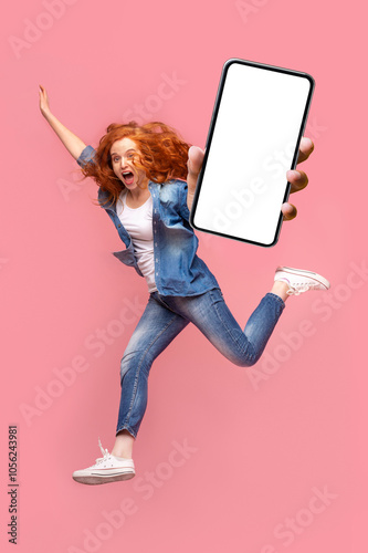 Happy young redhead woman jumping up, showing smartphone with empty screen, enjoying new application for mobile phone, full size photo. Pink studio background, creative image