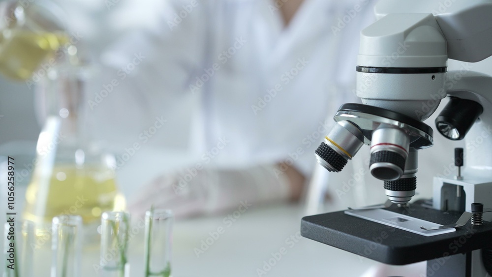 A scientist, wearing a lab coat and white protective gloves, is pouring a yellow oily liquid from one beaker to another near microscope in laboratory, close up. Medicine and science concept A scientist, wearing a lab coat and white protective gloves, is pouring a yellow oily liquid from one beaker to another near microscope in laboratory, close up. Medicine and science concept