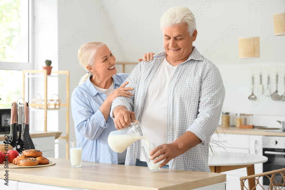Happy senior couple with tasty buns pouring milk from jug into glass in kitchen Happy senior couple with tasty buns pouring milk from jug into glass in kitchen