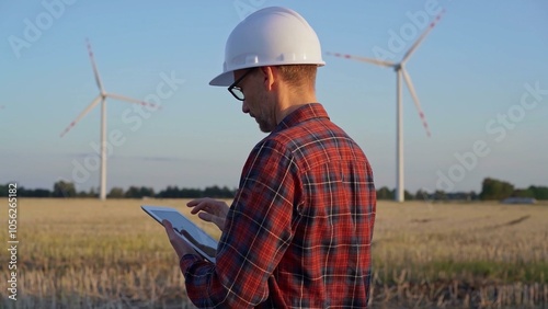 Man engineer at work is taking notes with a tablet computer in a field with wind turbines, as the sun sets. Clean energy concept