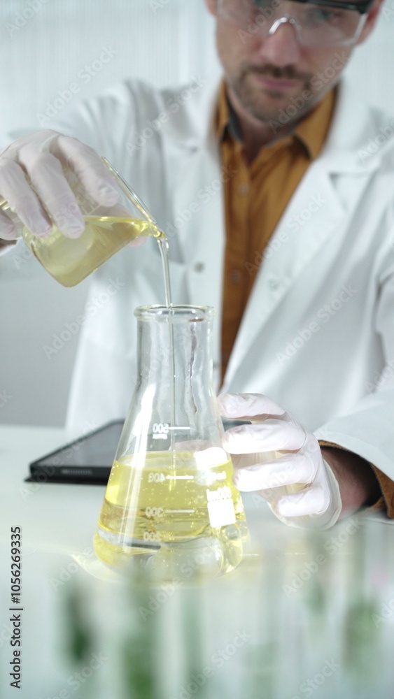 Man scientist with lab coat, white gloves, and protective glasses, is pouring a yellow oily liquid from one beaker to another near microscope in a laboratory, vertical close-up view Man scientist with lab coat, white gloves, and protective glasses, is pouring a yellow oily liquid from one beaker to another near microscope in a laboratory, vertical close-up view