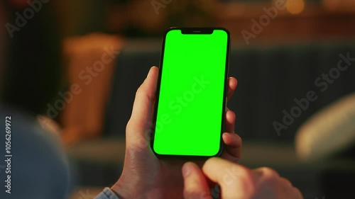 Man's hands holding smartphone with green screen in vertical position on blurred background of living room with sofa. Unrecognizable bearded hipster scrolling news feed. Indoors