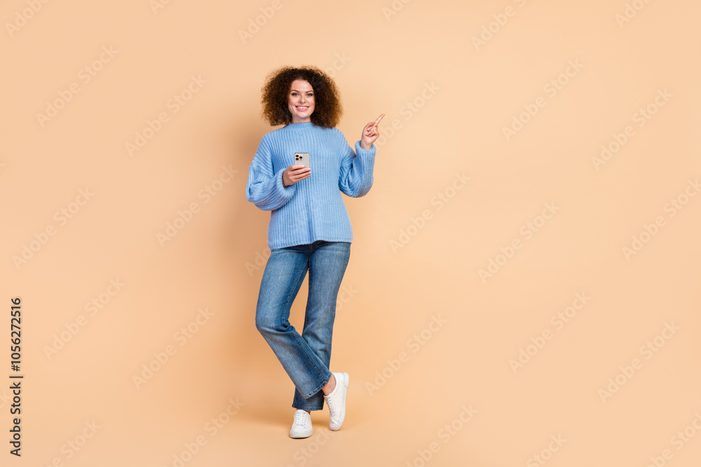 Photo of young blogger curly hair latin woman wearing blue knitted sweater with smartphone point finger website isolated on beige color background