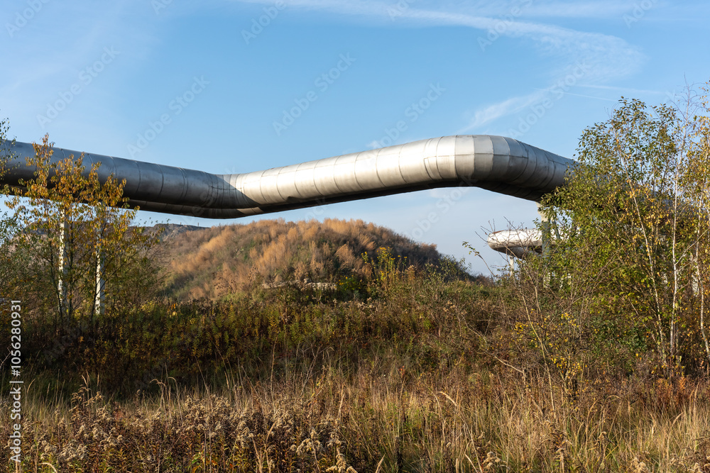 Elevated metal pipeline crossing over natural landscape with grass and trees on clear autumn day. Concept of industrial infrastructure coexisting with nature. Elevated metal pipeline crossing over natural landscape with grass and trees on clear autumn day. Concept of industrial infrastructure coexisting with nature.