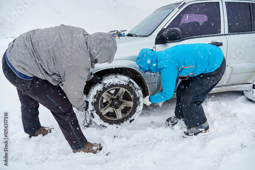 Travel in wintertime, pair of unrecognizable friends, man and woman, putting chains on the wheels of an SUV vehicle, in order to be able to move forward over the snow on a mountain road.