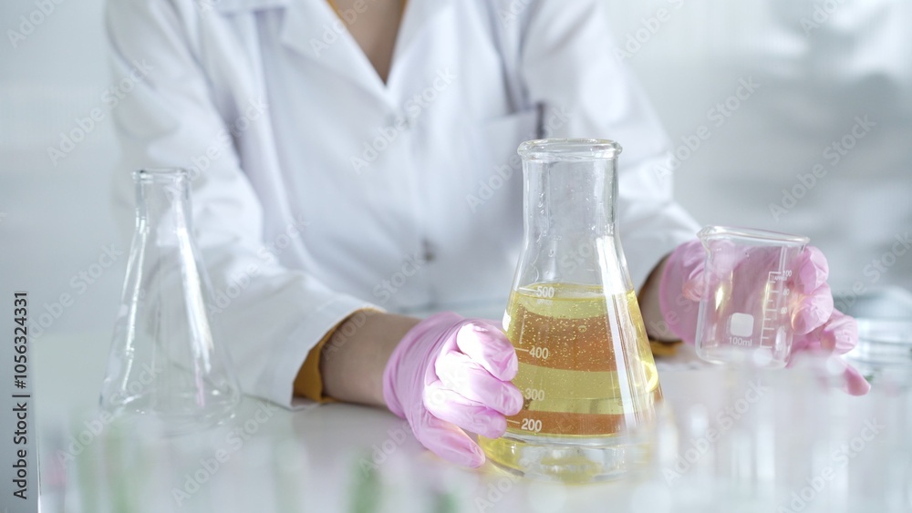 A scientist, wearing a lab coat and pink protective gloves, is pouring a yellow oily liquid from one beaker to another in laboratory, close up. Medicine and science A scientist, wearing a lab coat and pink protective gloves, is pouring a yellow oily liquid from one beaker to another in laboratory, close up. Medicine and science