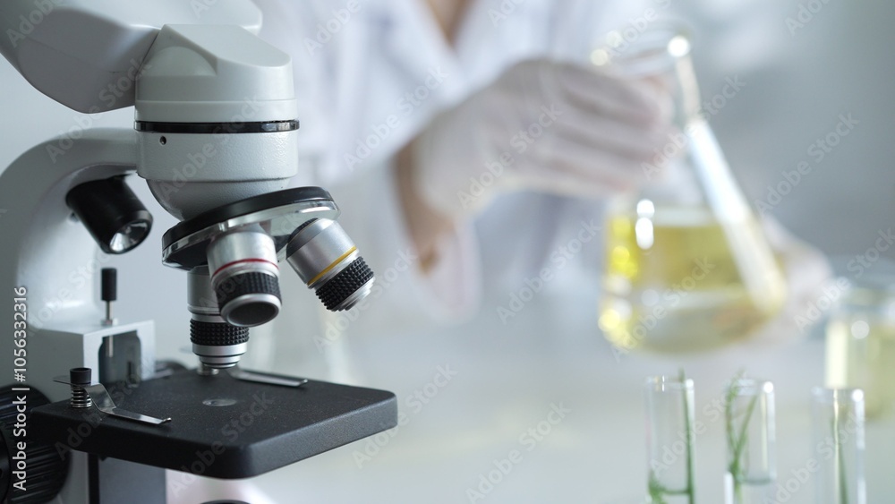 Microscope is near a scientist, wearing a lab coat and white protective gloves, and holding beaker with a yellow oily liquid in laboratory setting, close up. Medicine and science concept