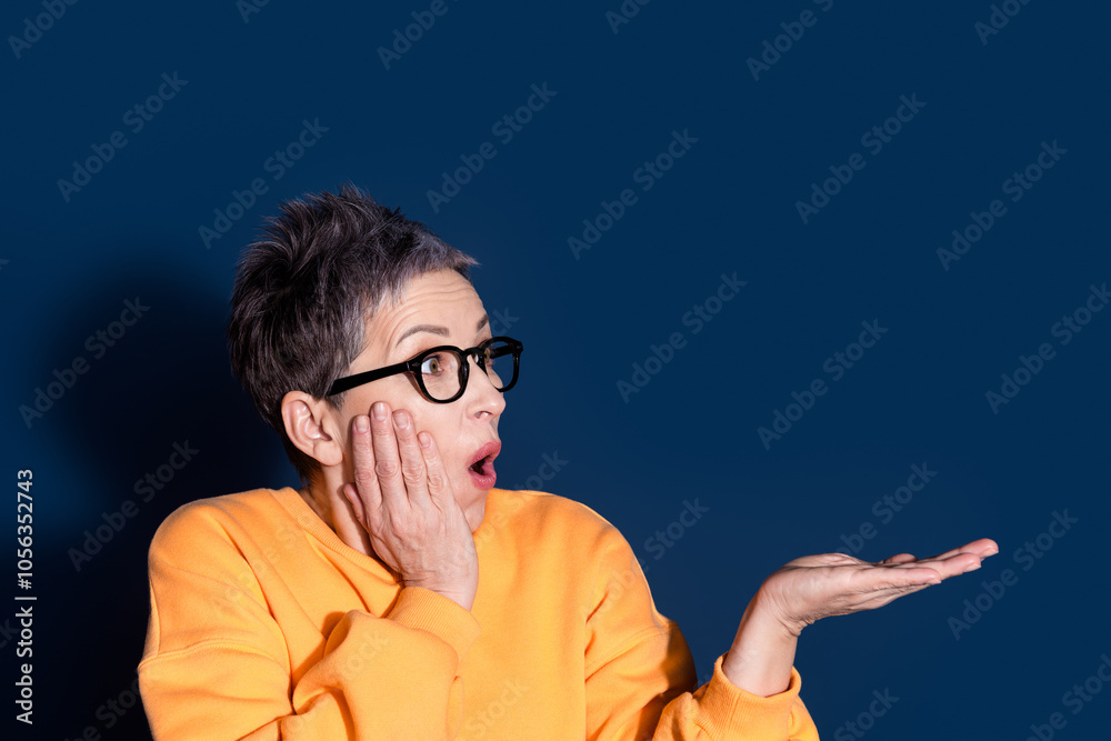 Photo portrait of pretty senior female look hold shocked empty space wear trendy yellow outfit isolated on dark blue color background Photo portrait of pretty senior female look hold shocked empty space wear trendy yellow outfit isolated on dark blue color background