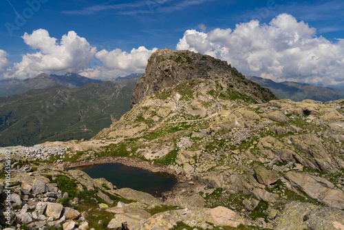 Picturesque view of rocky mountain landscape, small alpine pond in foreground. Rugged terrain and distant green valleys against blue sky with scattered clouds, capturing serene beauty of wilderness