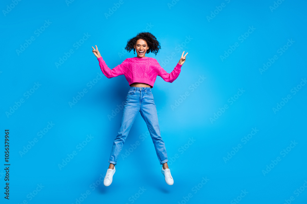 Full size photo of pretty young girl jumping show v-sign wear trendy pink outfit isolated on blue color background Full size photo of pretty young girl jumping show v-sign wear trendy pink outfit isolated on blue color background