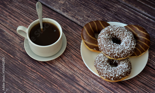 A cup of hot black coffee on a saucer with a spoon and a plate with several chocolate glazed donuts on a dark wooden table.